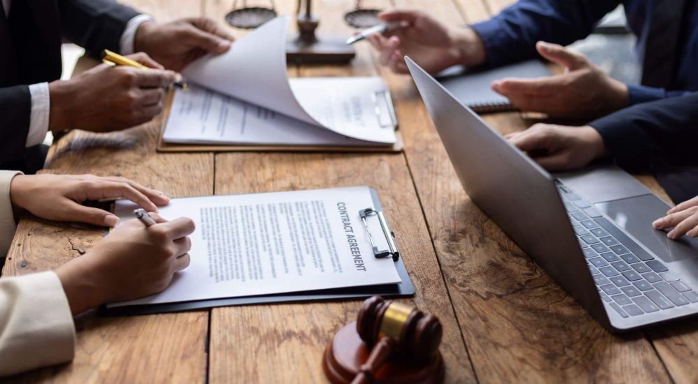 a group of people sitting around a wooden table and working with multiple documents and laptops