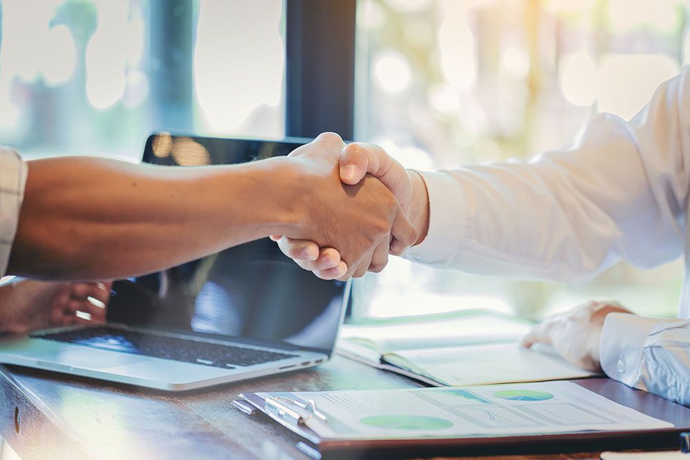 Handshake over a desk with a laptop and documents, symbolizing a successful business partnership.