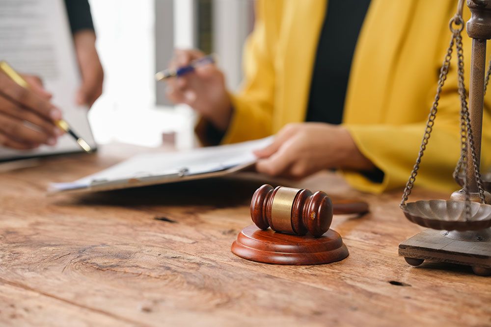 Legal consultation scene with gavel on table, people reviewing documents, and scales of justice in the background.