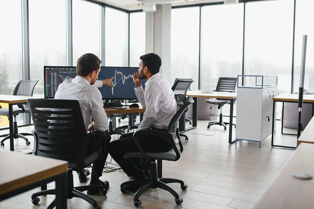 Two businessmen analyzing financial graphs on a computer in a modern office setting.