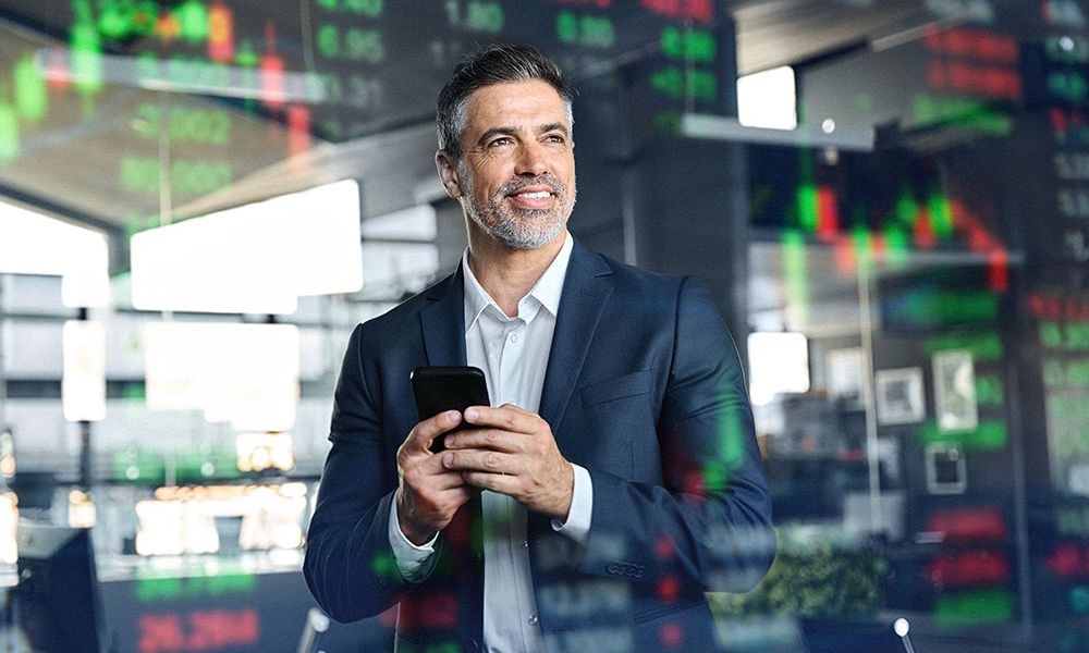 Businessman in office smiling while looking at smartphone, stock market data projected in the background.