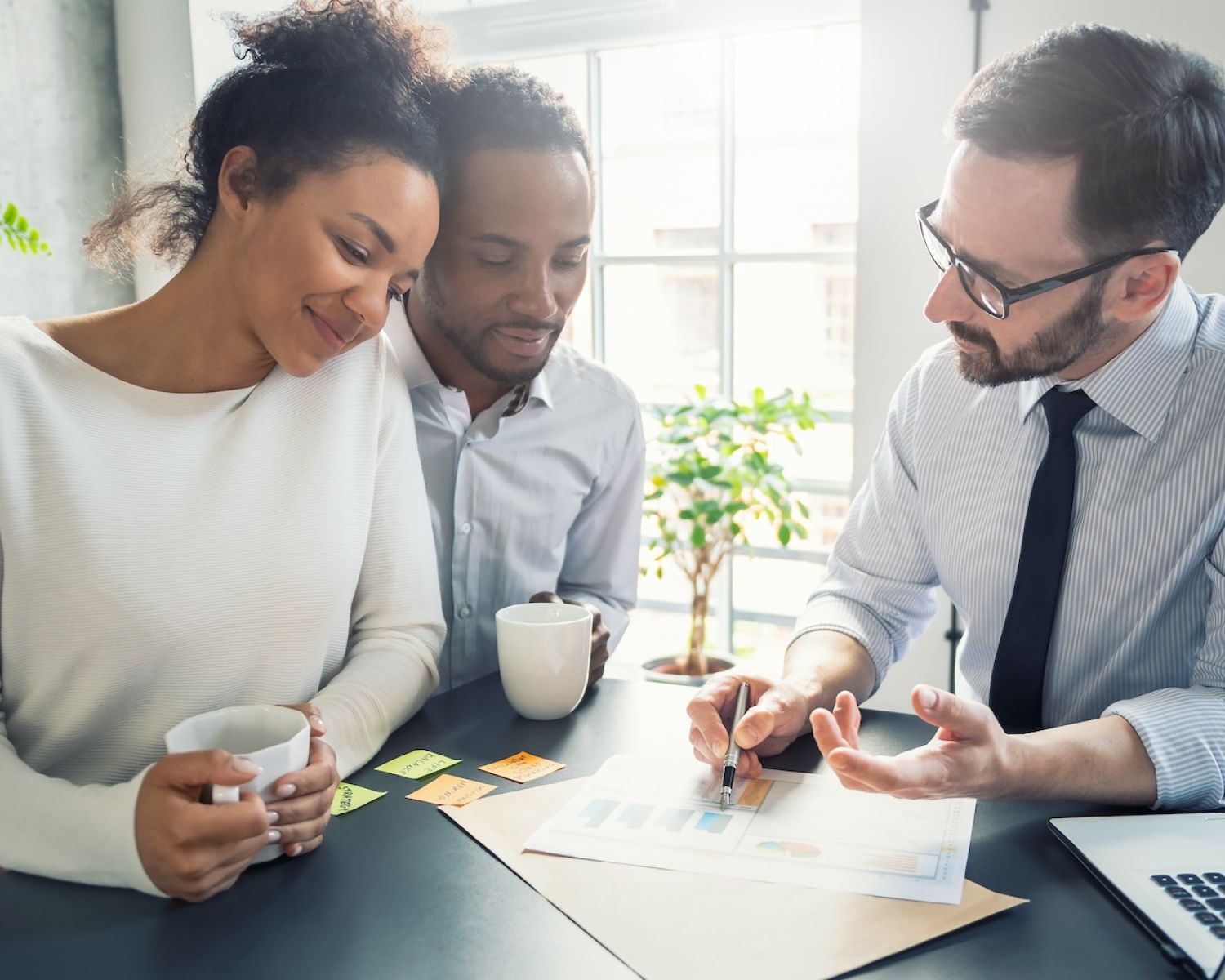 Couple consulting with a financial advisor, discussing documents with charts at a desk, coffee cups in hand.