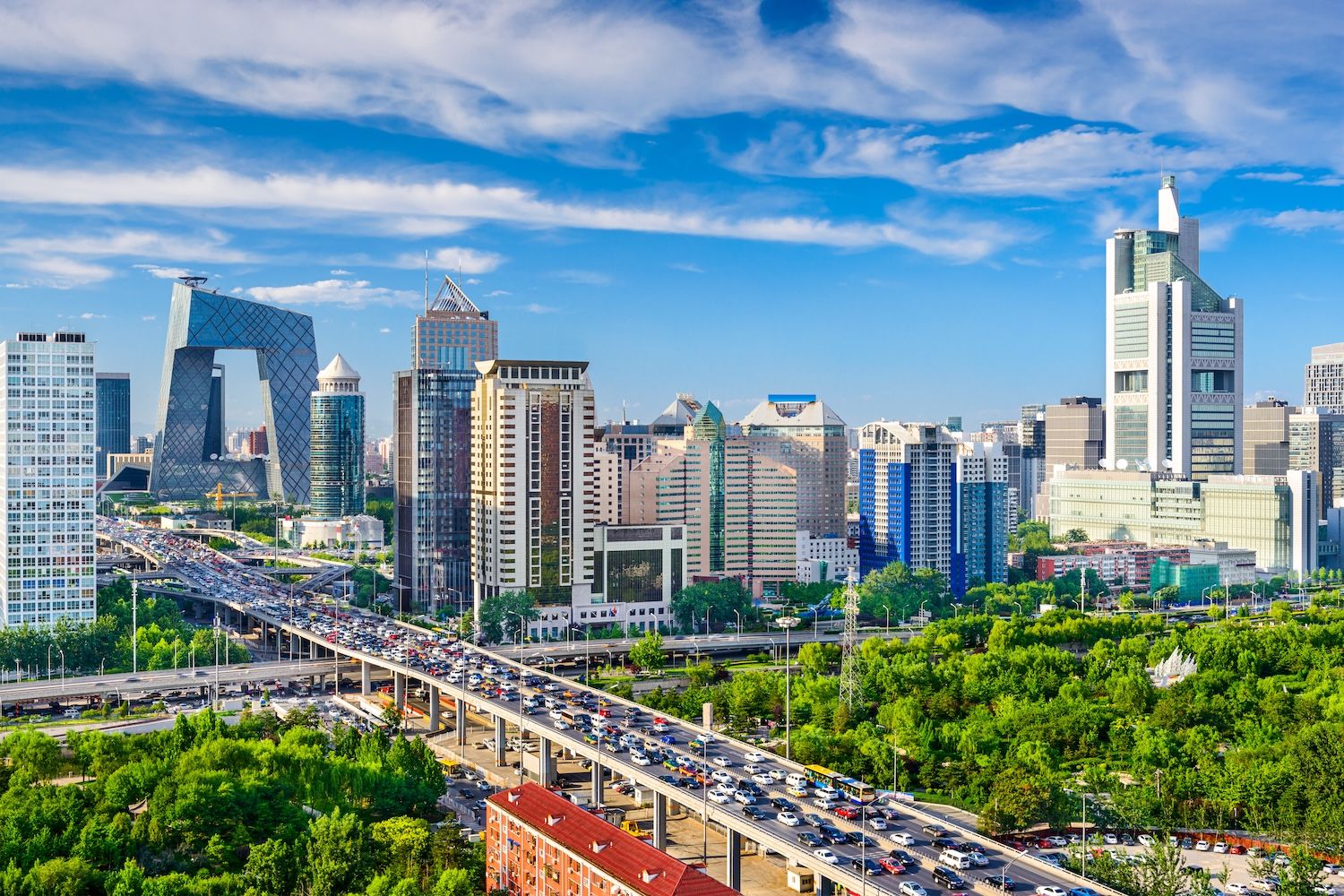 Beijing cityscape with modern skyscrapers, lush greenery, and a busy highway under a vibrant blue sky.