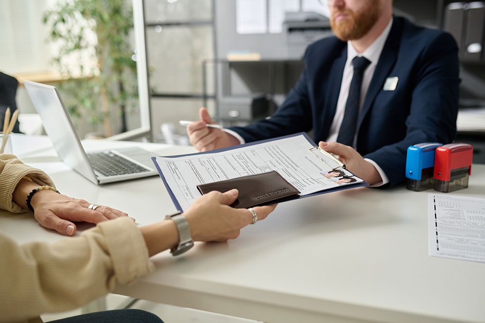 Person handing over documents to a business professional across a desk with a laptop, stamps, and papers.
