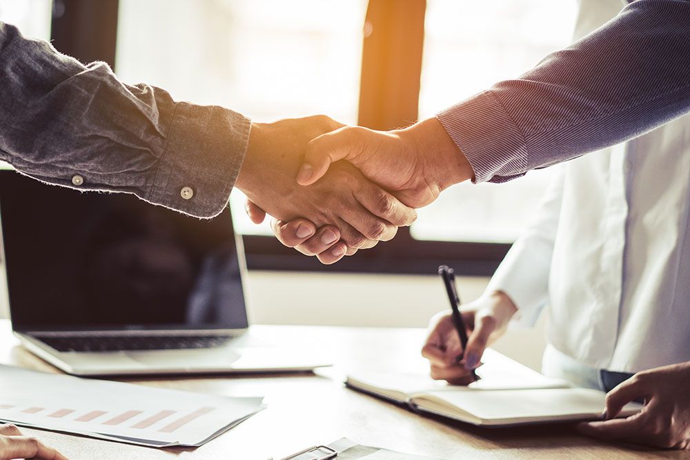 Business handshake in office, agreement concept, two people shaking hands near a laptop and documents on a desk.