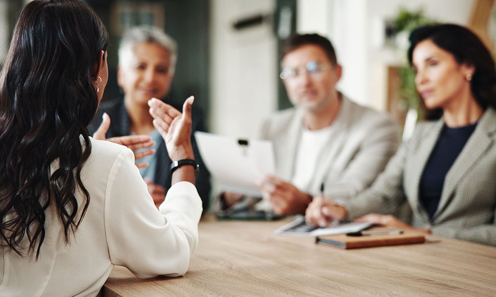 Woman in white shirt at meeting table discussing with three colleagues in office setting.