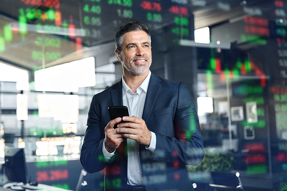 Businessman in office smiling while looking at smartphone, stock market data projected in the background.
