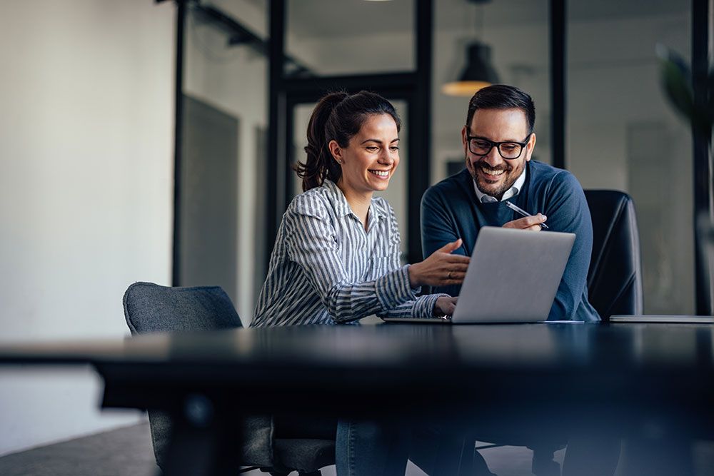 Two colleagues smiling while working on a laptop in a modern office setting.