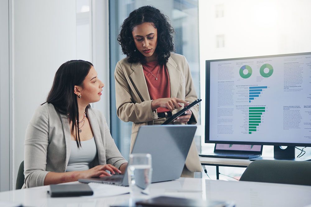 Two businesswomen discussing data on a tablet, with charts displayed on a computer screen in a modern office setting.