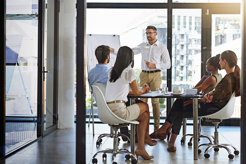 Business meeting presentation with diverse team in a bright office, featuring a man pointing at a whiteboard.