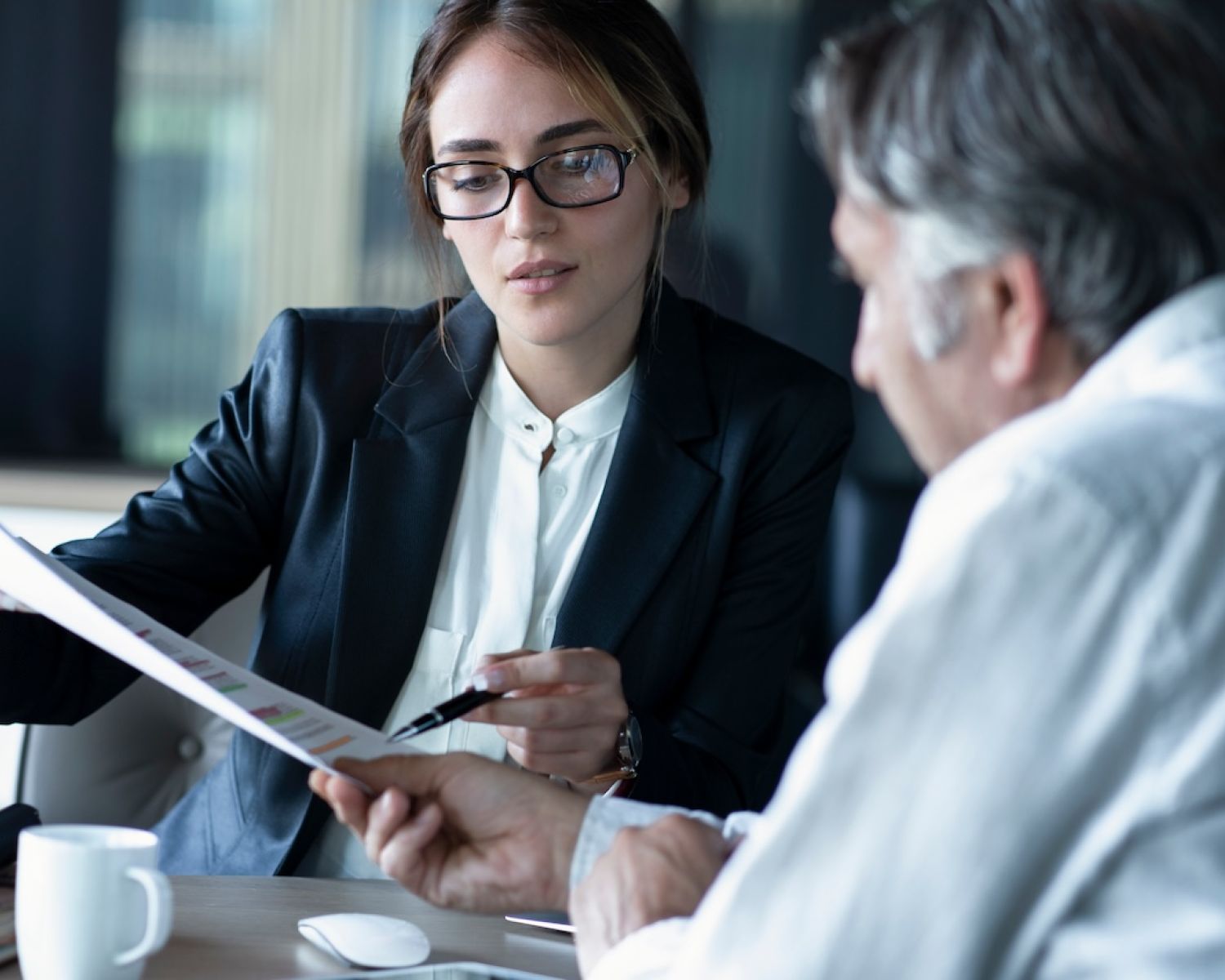 Business meeting: woman in glasses discussing a document with a colleague across a desk in an office setting.
