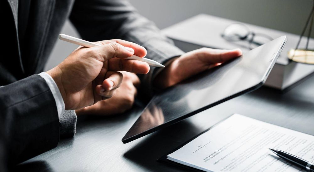 Hands hold a tablet while another hand points with a stylus. A document with a pen lies on a dark table, near a pair of glasses in a professional setting.
