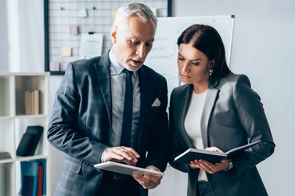 Business professionals reviewing documents on a tablet and notebook in an office setting.