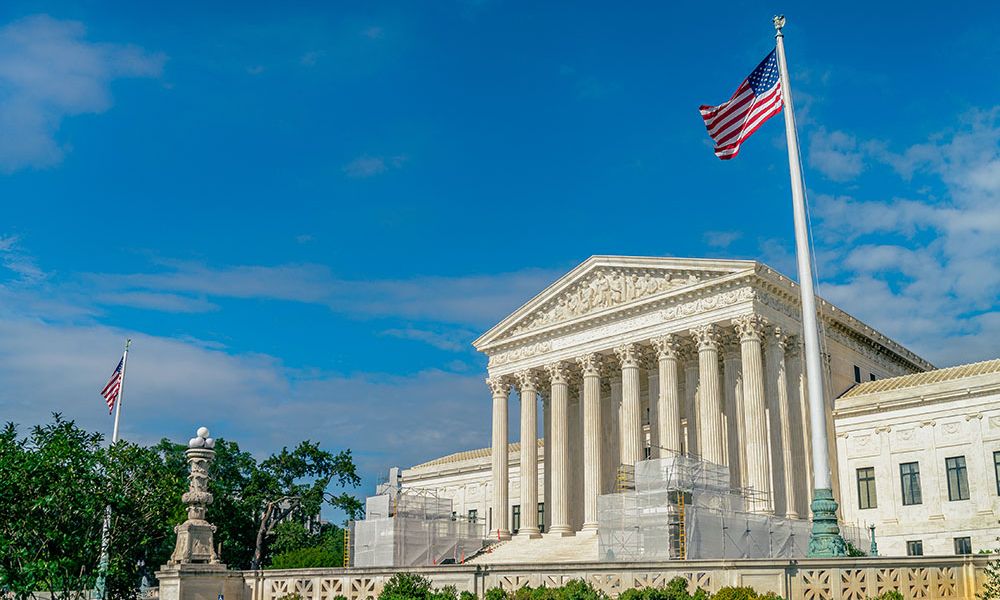 U.S. Supreme Court building with American flag under clear blue sky, showcasing neoclassical architecture.