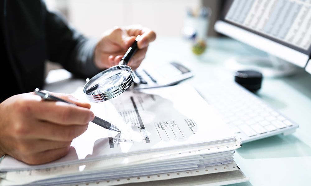Person examining financial documents with magnifying glass at desk.