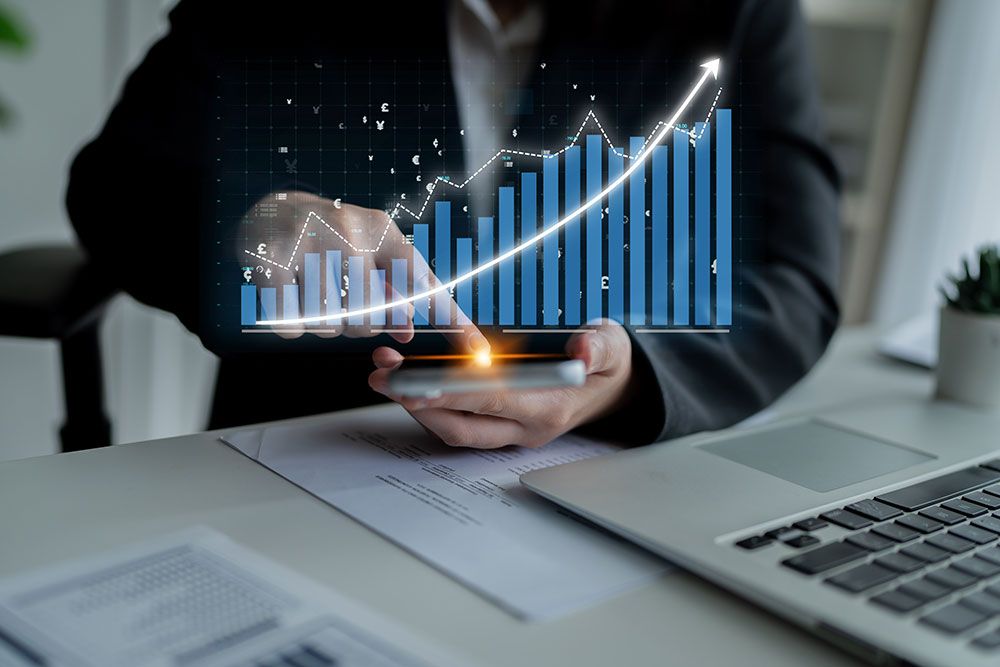 Person analyzes digital financial chart with growth trends on a smartphone, sitting at a desk with a laptop.