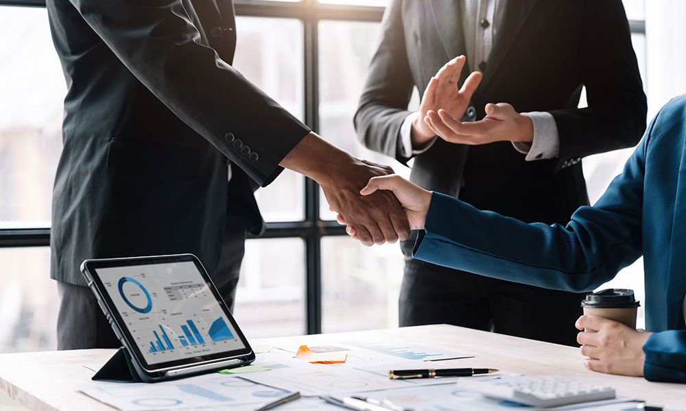 Two professionals shake hands across a desk, signifying a business agreement or partnership.