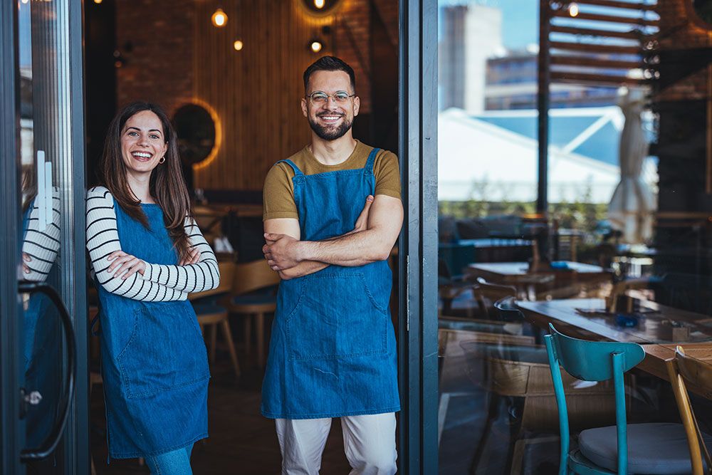 Two smiling cafe owners in blue aprons stand at the entrance of their cozy, modern coffee shop.