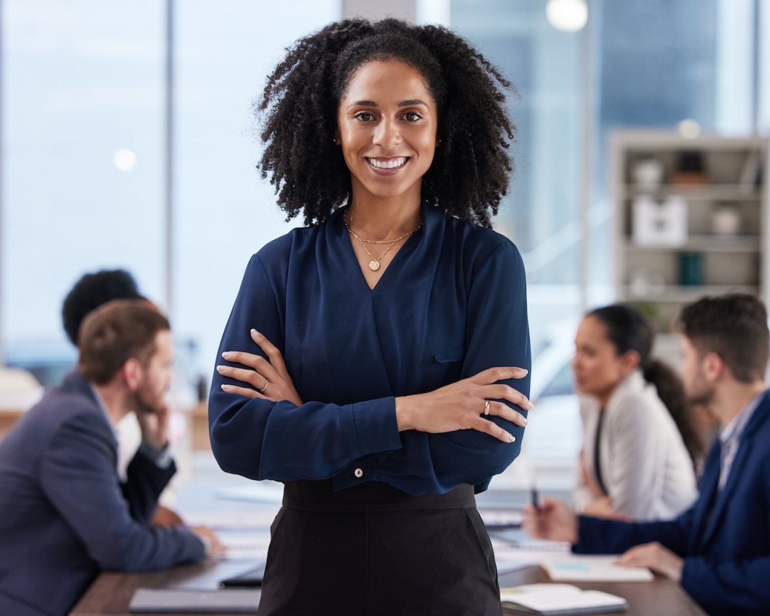 Confident businesswoman smiling in front of team meeting, wearing blue blouse, professional office setting.
