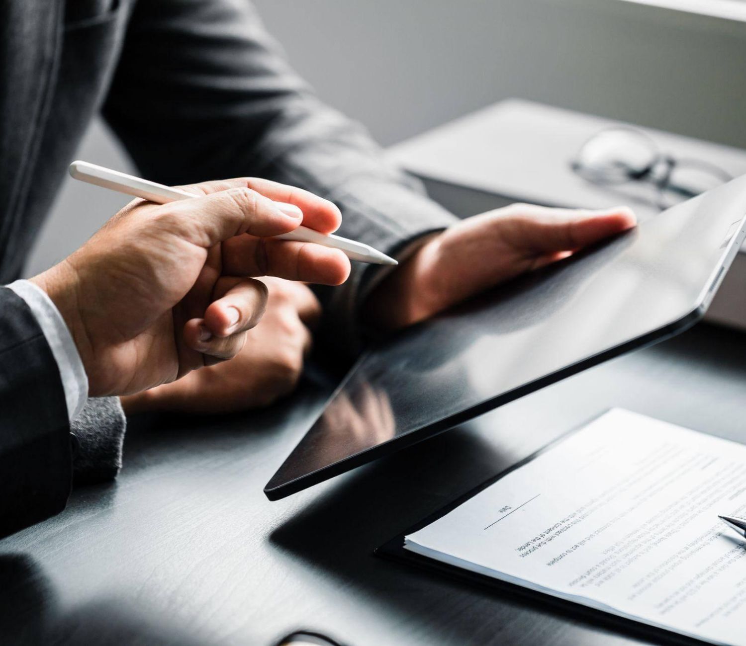 Hands hold a tablet while another hand points with a stylus. A document with a pen lies on a dark table, near a pair of glasses in a professional setting.