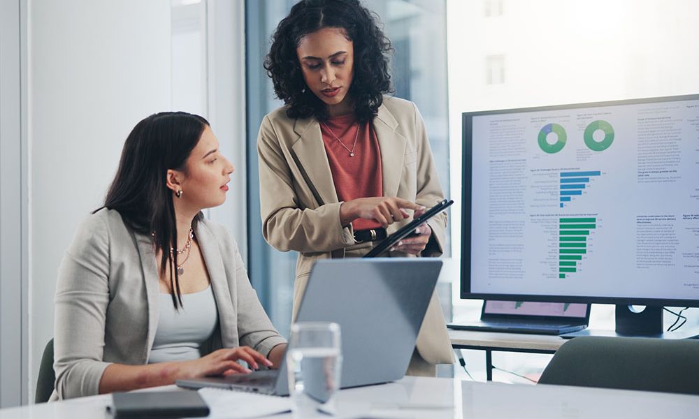 Two businesswomen discussing data on a tablet, with charts displayed on a computer screen in a modern office setting.