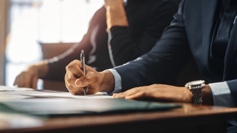 Person in a suit writing with a pen at a desk, close-up of hand, professional setting.