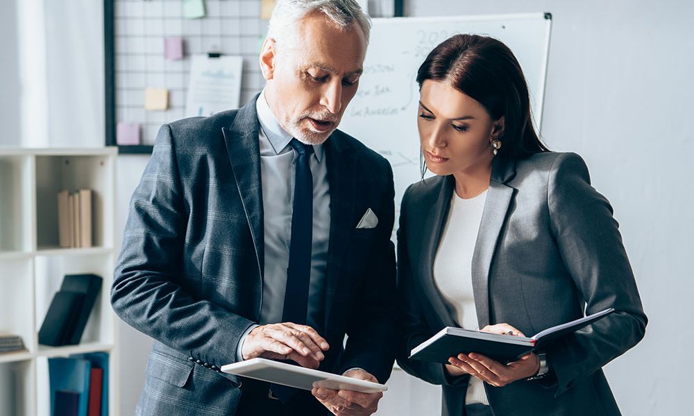 Business professionals reviewing documents on a tablet and notebook in an office setting.