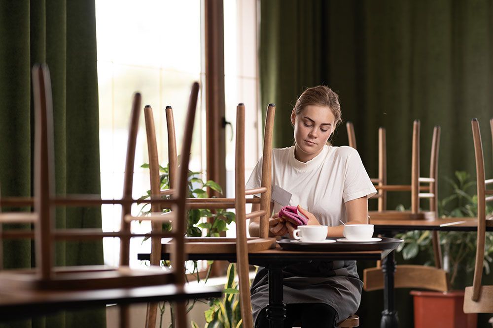 Woman seated in empty café with stacked chairs, using phone and drinking coffee.
