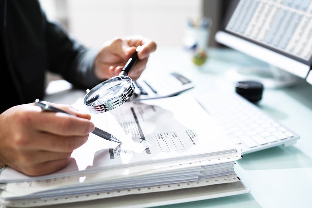 Person examining financial documents with magnifying glass at desk.