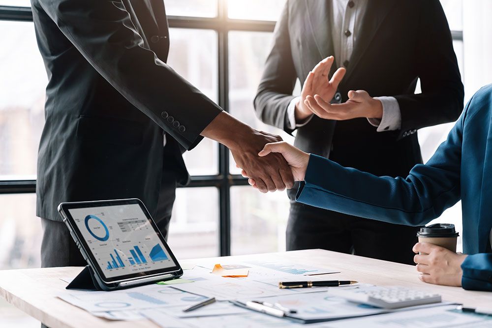 Two professionals shake hands across a desk, signifying a business agreement or partnership.