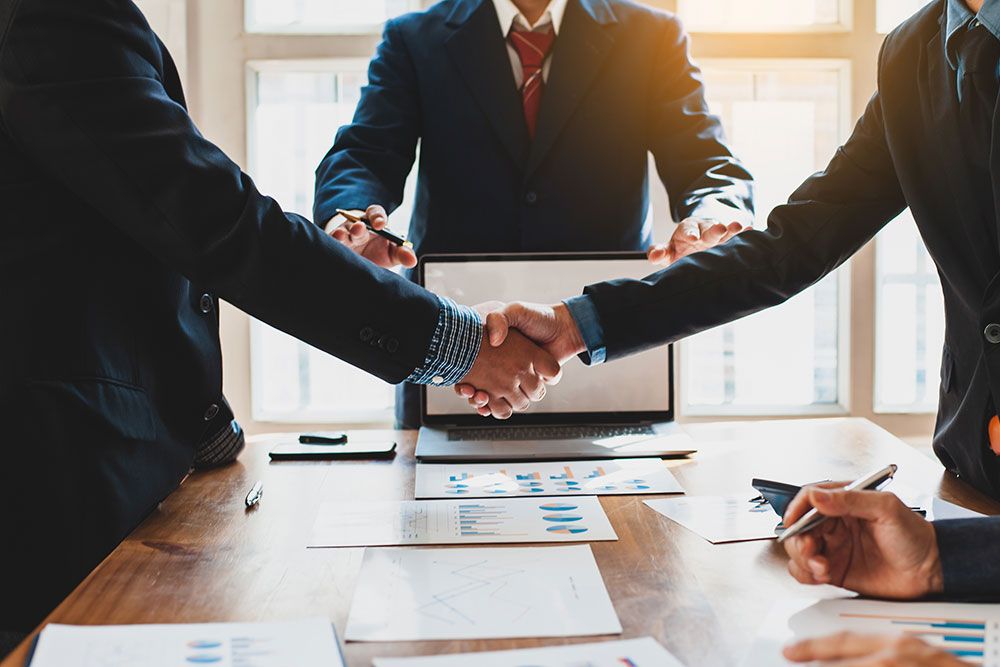 Business professionals shaking hands over a meeting table with documents and a laptop, symbolizing agreement and partnership.