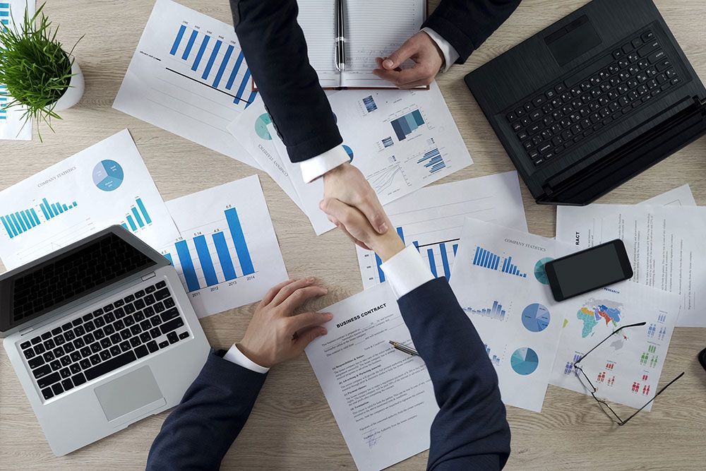 Two businesspeople shaking hands over a contract and financial charts on a desk with laptops and a smartphone.