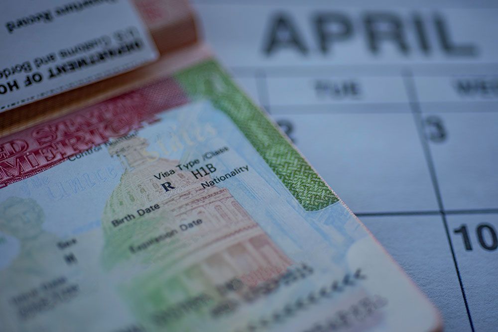 Close-up of an H-1B visa on a passport with a blurred calendar showing the month of April in the background.