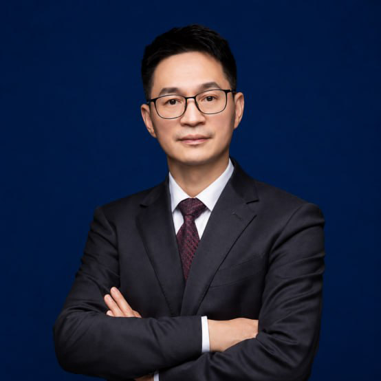 정장을 입고 곱슬머리를 한 전문직 남성이 회사 환경에서 미소를 짓고 있다. 배경은 흐릿하게 처리되어 있다.