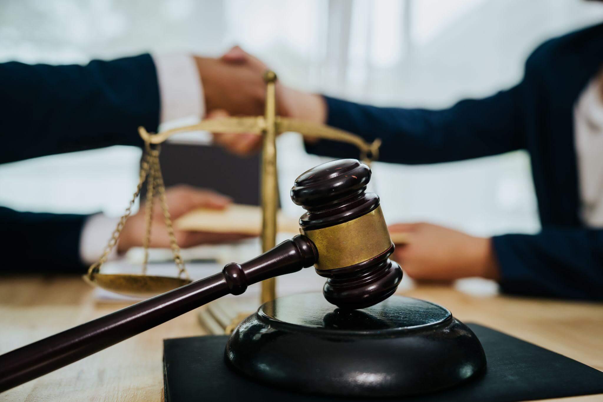 A judge's gavel rests on a desk beside scales of justice. In the background, two people in suits shake hands, symbolizing agreement or resolution in a formal setting.