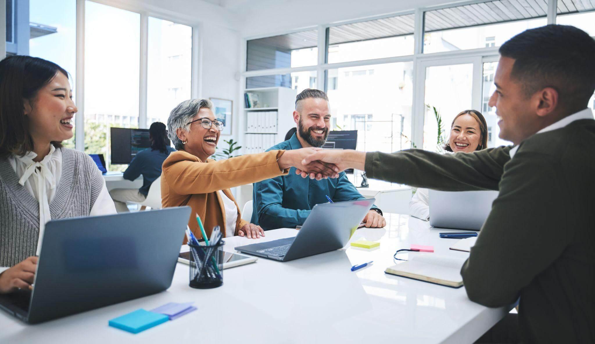a group of people sitting around a table shaking hands; they have their laptops open and are working in a big, bright office